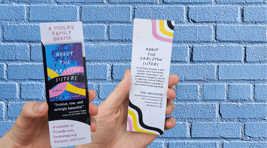 A hand holds up two promotional bookmarks in front of a brick wall.